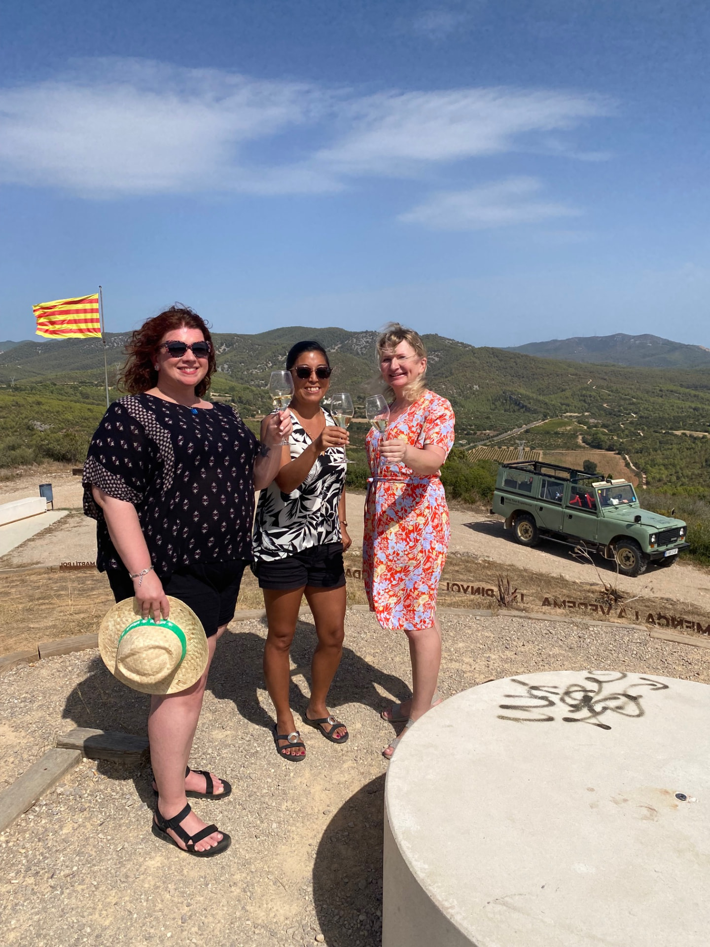 Three guests toasting Cava at a hilltop viewpoint with the Catalan flag and a vintage Land Rover, hosted by Alexandra Kalinina, WSET Level 3 certified Cava expert and private wine guide